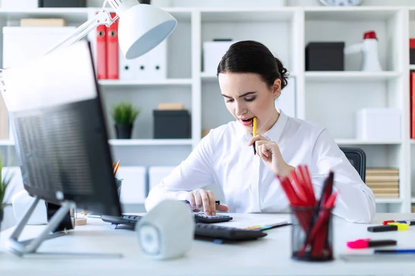 Een jong meisje in het Bureau houdt een pen in haar mond en werkt met een rekenmachine, documenten en een computer. — Stockfoto