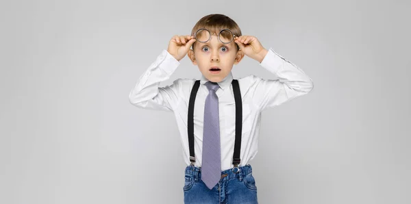 Ein charmanter Junge in weißem Hemd, Hosenträgern, Krawatte und heller Jeans steht vor grauem Hintergrund. Der Junge hält sich an seine Brille — Stockfoto