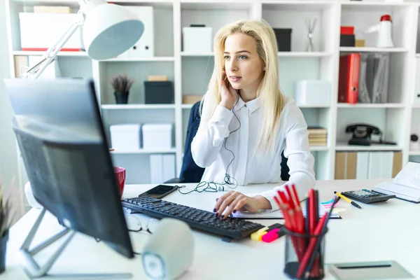Uma jovem no escritório senta-se em uma mesa conversando ao telefone através de um fone de ouvido e trabalhando no computador . — Fotografia de Stock