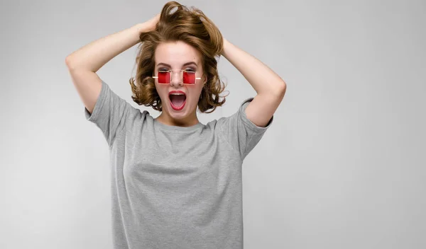 Charmantes junges Mädchen in einem grauen T-Shirt vor grauem Hintergrund. Mädchen mit roter quadratischer Brille. — Stockfoto