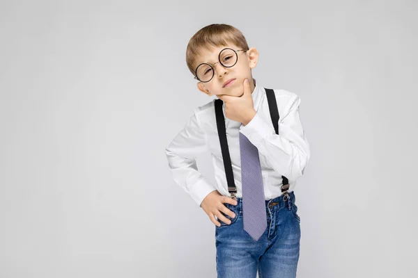 Un niño encantador con una camisa blanca, tirantes, corbata y pantalones vaqueros ligeros se levanta sobre un fondo gris. un chico con gafas está sosteniendo una mano cerca de su cara — Foto de Stock