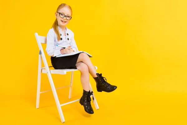 Flicka med rött hår på gul bakgrund. En charmig tjej i transparent glas sitter på en vit stol och gör anteckningar i en anteckningsbok. — Stockfoto