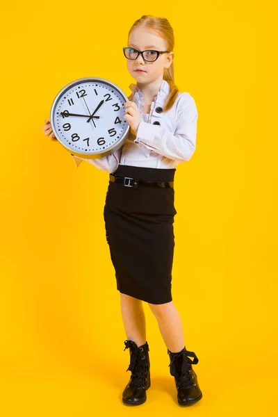 Mädchen mit roten Haaren auf gelbem Hintergrund. ein charmantes Mädchen mit durchsichtiger Brille hält eine Uhr. — Stockfoto