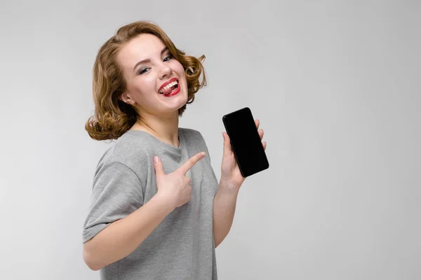 Charmantes junges Mädchen in einem grauen T-Shirt vor grauem Hintergrund. ein Mädchen hält ein Handy in der Hand — Stockfoto