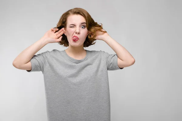 Charming young girl in a gray T-shirt on a gray background. Girl showing tongue — Stock Photo, Image