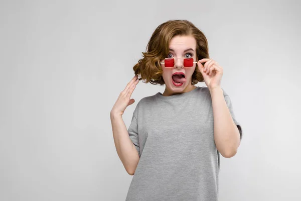 Encantadora joven con una camiseta gris sobre un fondo gris. Chica en gafas de ojos cuadrados rojos . — Foto de Stock