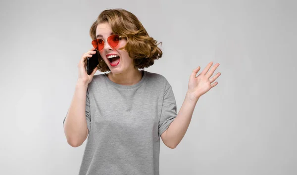 Encantadora joven con una camiseta gris sobre un fondo gris. Chica en gafas rojas con teléfono — Foto de Stock