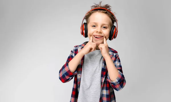 Ένα όμορφο αγόρι σε ένα καρό πουκάμισο, γκρι πουκάμισο και τζιν στέκεται σε γκρι φόντο. Ένα αγόρι στο κόκκινο ακουστικά. Το αγόρι τεντώνει τα δάχτυλά του με ένα χαμόγελο. — Φωτογραφία Αρχείου