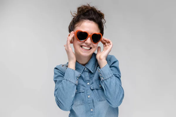 Junges brünettes Mädchen mit Brille in Form eines Herzens. Die Haare sind zu einem Dutt zusammengebunden. das Mädchen justiert seine Brille. — Stockfoto