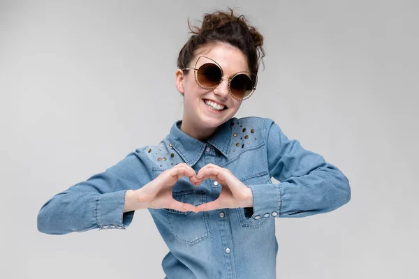 Young brunette girl in black glasses. Cat glasses. The hair is gathered in a bun. The girl shows the hands of the heart. — Stock Photo, Image
