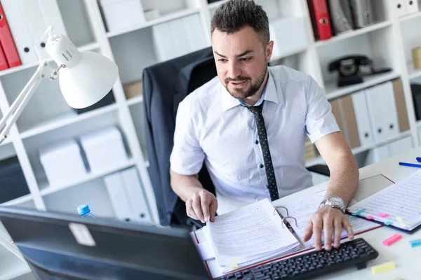 Bir adam belgeleri ve bir bilgisayar ile çalışma ofisinde, bir masada oturuyor.