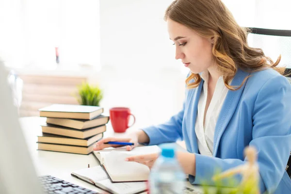 Mooi jong meisje zitten aan de balie in kantoor, het bezit van een pen in haar hand en het lezen van een boek. — Stockfoto