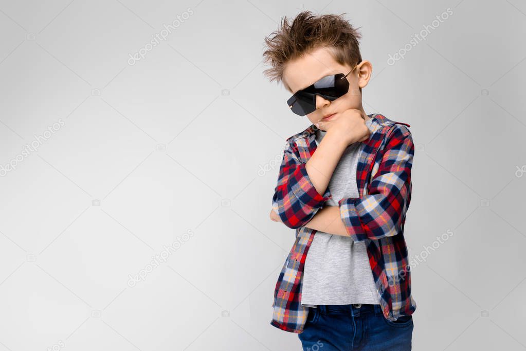 A handsome boy in a plaid shirt, gray shirt and jeans stands on a gray background. The boy in black sunglasses. The boy holds a hand to his chin