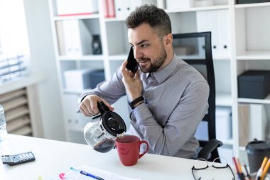 Ofiste genç bir adam üstünde belgili tanımlık telefon ve dökme kahve bir fincan içine konuşmak bir masada oturur.