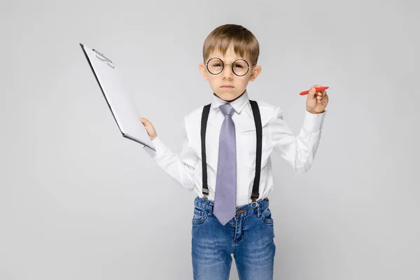 Ein charmanter Junge in weißem Hemd, Hosenträgern, Krawatte und heller Jeans steht vor grauem Hintergrund. der Junge hält einen Stift und Blätter für Notizen — Stockfoto