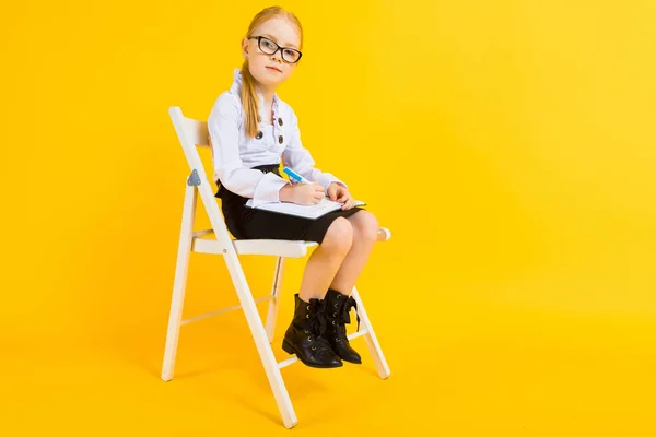 Mädchen mit roten Haaren auf gelbem Hintergrund. ein charmantes Mädchen mit durchsichtiger Brille sitzt auf einem weißen Stuhl und macht Notizen in einem Notizbuch. — Stockfoto