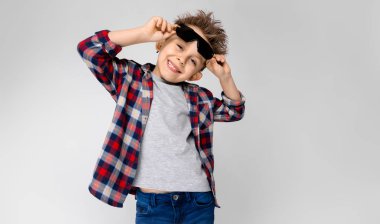 Yakışıklı bir çocuk bir ekose gömlek, gri gömlek ve pantolon gri bir arka plan üzerinde duruyor. Siyah güneş gözlüklü çocuk. Çocuk gözlüğünü tutuyor.