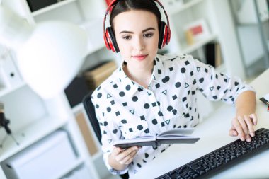 Güzel bir genç kız kulaklık ofiste bir masada oturuyor, onun elinde bir defter tutan ve metin üstünde belgili tanımlık klavye türleri.
