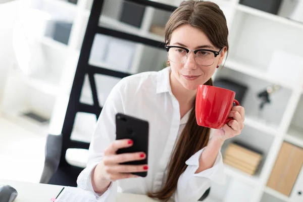 Una joven está sentada en una mesa en la oficina, sosteniendo un teléfono y una taza roja en su mano . — Foto de Stock