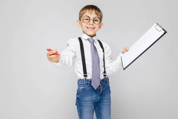Очаровательный мальчик в белой рубашке, подтяжках, галстуке и легких джинсах стоит на сером фоне. Мальчик держит ручку и простыни для заметок — стоковое фото