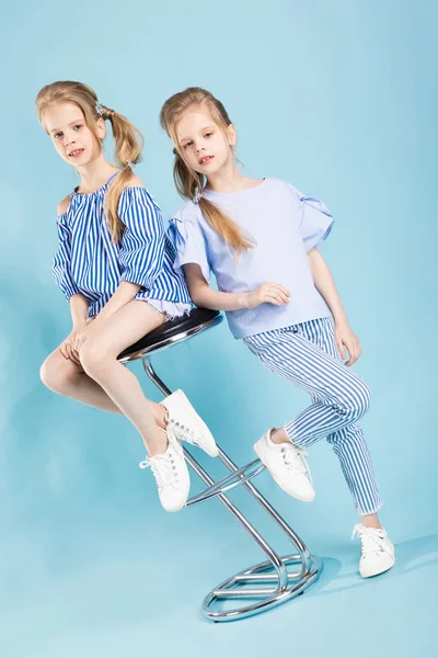 Mädchen-Zwillinge in hellblauer Kleidung posieren in der Nähe eines Barhockers auf blauem Hintergrund. — Stockfoto