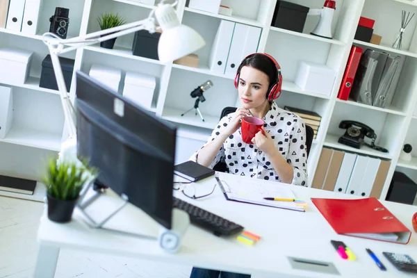 Uma jovem se senta em fones de ouvido em uma mesa no escritório, segura uma xícara vermelha em suas mãos e olha para o monitor . — Fotografia de Stock