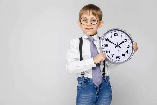 Ein charmanter Junge in weißem Hemd, Hosenträgern, Krawatte und heller Jeans steht vor grauem Hintergrund. der Junge hält eine Armbanduhr — Stockfoto