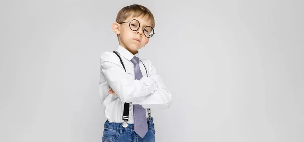 Okouzlující chlapec v bílé košili, šle, kravatu a lehké džíny stojí na šedém pozadí. Ten založil ruce na prsou — Stock fotografie