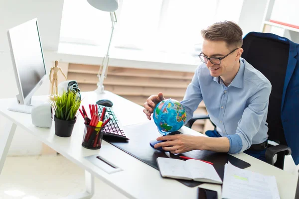 A young man sits in the office at a computer desk and holds a globe in his hands.