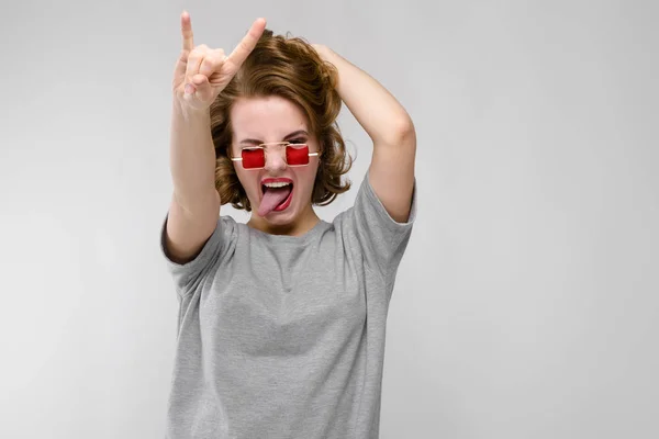 Charmantes junges Mädchen in einem grauen T-Shirt vor grauem Hintergrund. Mädchen mit roter eckiger Brille. Das Mädchen zeigt eine Rockerziege — Stockfoto