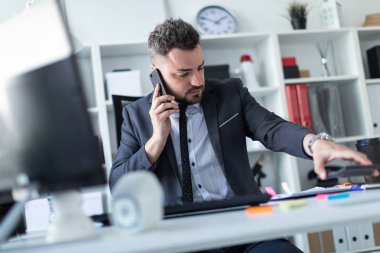 Bir adam masada ofiste oturur, telefonda konuşuyor, belgeleriyle çalışır ve bir kenara koyar hesap makinesi.