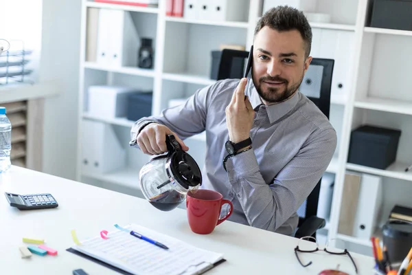 Un joven en la oficina se sienta en una mesa, hablando por teléfono y sirviendo café en una taza . — Foto de Stock