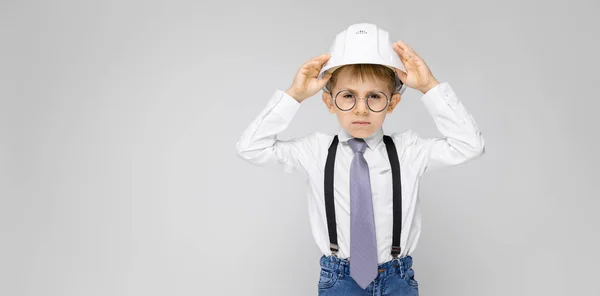 Ein charmanter Junge in weißem Hemd, Hosenträgern, Krawatte und heller Jeans steht vor grauem Hintergrund. Junge mit weißem Helm — Stockfoto