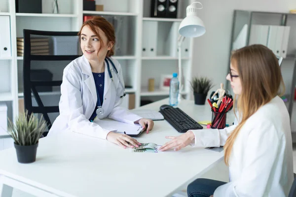 Ein schönes junges Mädchen in weißem Gewand sitzt am Schreibtisch im Büro und kommuniziert mit dem Gesprächspartner. Der Gesprächspartner gibt dem Mädchen Geld. — Stockfoto