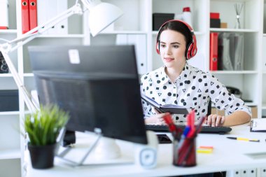Güzel genç kız kulaklık Office masada oturur, onun elinde bir defter tutan ve klavyede yazdırır. Alan, kız odak derinliği ile fotoğraf.
