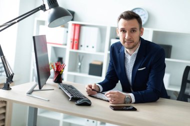 Genç bir adam ofiste bir tabloda yakınındaki duruyor, bir kalem elinde tutuyor ve belgeler ve bir bilgisayar ile çalışır.