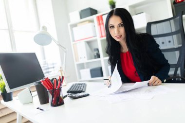 Güzel genç kız ofiste belgelerle masada çalışıyor ve bir kalem elinde tutuyor.