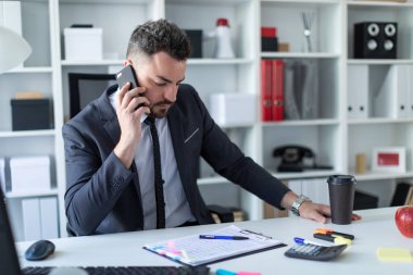 Bir adam ofiste masa başında oturan, telefonda konuşurken ve bir bardak kahve holding.