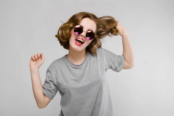 Charmantes junges Mädchen in einem grauen T-Shirt vor grauem Hintergrund. fröhliches Mädchen mit runder Brille hält eine Hand voller Haare — Stockfoto