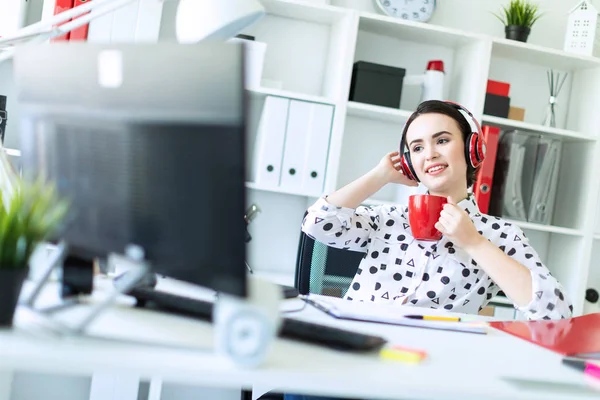 Een jong meisje zit in de koptelefoon aan een tafel in het kantoor, houdt een rode kop in haar handen en kijkt naar de monitor. — Stockfoto