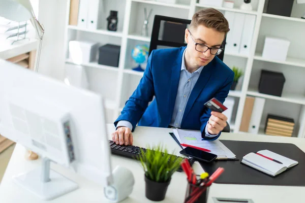 Un joven está sentado en una mesa en la oficina, sosteniendo una tarjeta bancaria en su mano y escribiendo en una computadora . — Foto de Stock