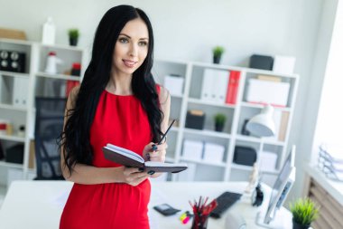 Kırmızı elbiseli güzel genç kız ofiste duran ve bir defter ve bir kalem tutan.