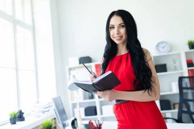Kırmızı elbiseli güzel genç kız ofiste duran ve bir defter ve bir kalem tutan.
