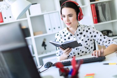 Güzel genç kız kulaklık Office masada oturur, onun elinde bir defter tutan ve klavyede yazdırır. Alan, kız odak derinliği ile fotoğraf.