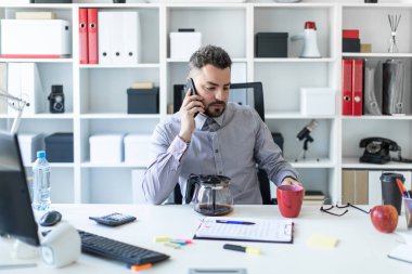 Ofiste genç bir adam telefonda konuşurken ve kırmızı bir fincan elinde tutan bir masada oturur.