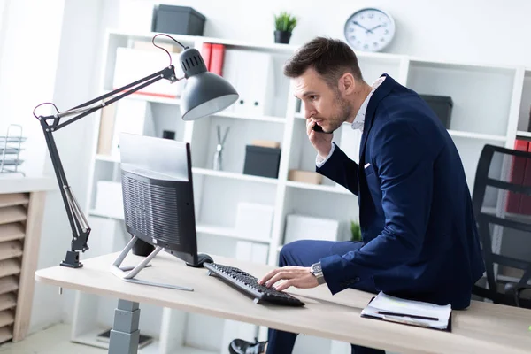 Молодой человек сидит в офисе на столе, разговаривает по телефону и работает с компьютером . — стоковое фото