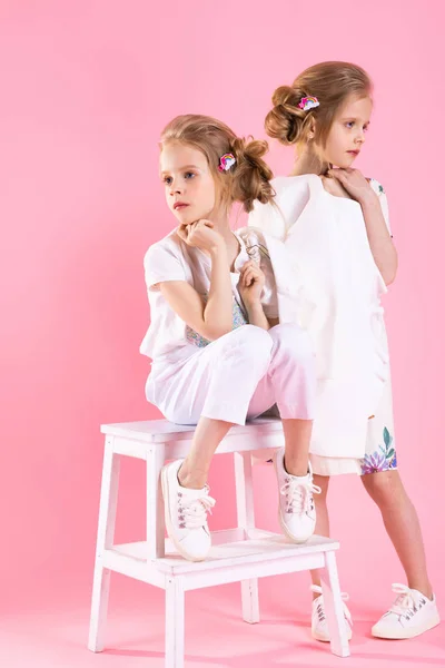Zwillinge Mädchen in hellen Kleidern posieren in der Nähe der Treppe mit zwei Schritten auf einem rosa Hintergrund. — Stockfoto