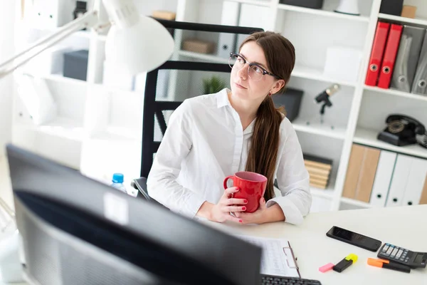 En ung flicka sitter vid ett bord på kontoret och innehar en röd kopp i händerna. — Stockfoto