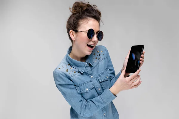 Young brunette girl in round glasses. Hairs are gathered in a bun. Girl with a black phone. The girl is looking at the phone. — Stock Photo, Image