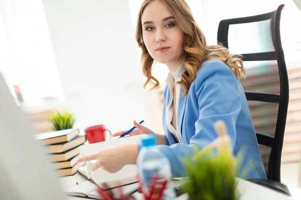 Mooi jong meisje zitten aan de balie in kantoor, het bezit van een pen in haar hand en het lezen van een boek. — Stockfoto